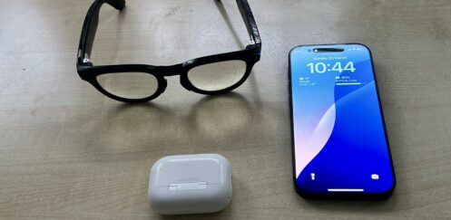 A pair of Ray-Ban Meta smart glasses next to an iPhone and AirPods, highlighting the interoperability gap with Apple’s AirPods, which have exclusive accessibility features