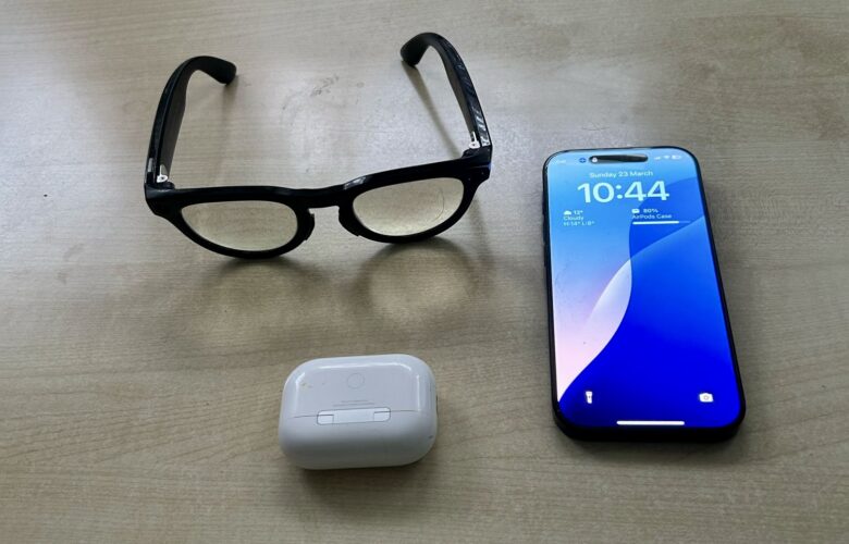 A pair of Ray-Ban Meta smart glasses next to an iPhone and AirPods, highlighting the interoperability gap with Apple’s AirPods, which have exclusive accessibility features