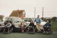 1990s family photo of Colin Hughes with siblings Ian and Lyn; three wheelchair-accessible Motability Chrysler Voyager vehicles behind them.