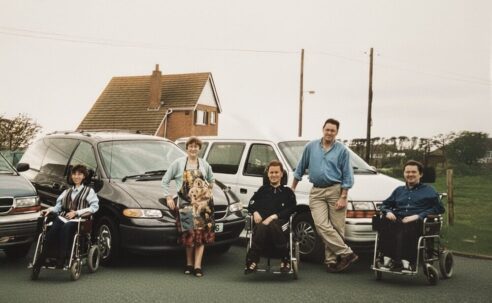 1990s family photo of Colin Hughes with siblings Ian and Lyn; three wheelchair-accessible Motability Chrysler Voyager vehicles behind them.