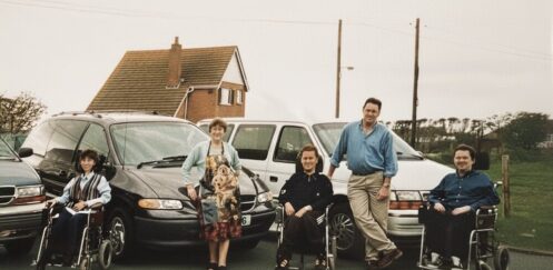 1990s family photo of Colin Hughes with siblings Ian and Lyn; three wheelchair-accessible Motability Chrysler Voyager vehicles behind them.