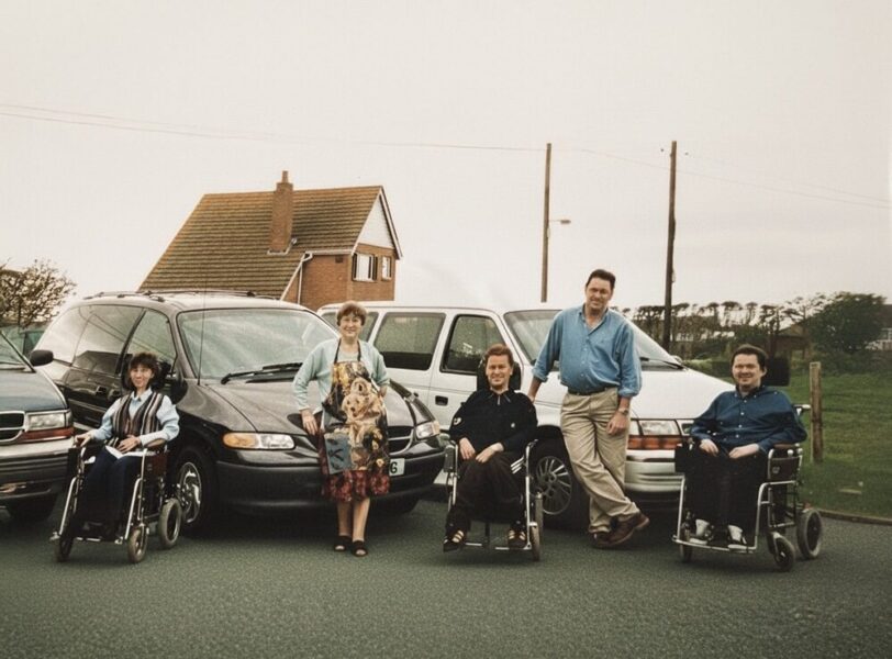 1990s family photo of Colin Hughes with siblings Ian and Lyn; three wheelchair-accessible Motability Chrysler Voyager vehicles behind them.