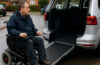 A wheelchair user beside an adapted Motability vehicle on a residential street, illustrating the impact of Motability cuts.