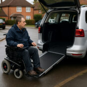 A wheelchair user beside an adapted Motability vehicle on a residential street, illustrating the impact of Motability cuts.