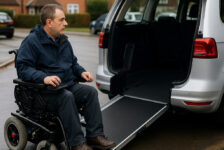 A wheelchair user beside an adapted Motability vehicle on a residential street, illustrating the impact of Motability cuts.