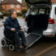 A wheelchair user beside an adapted Motability vehicle on a residential street, illustrating the impact of Motability cuts.