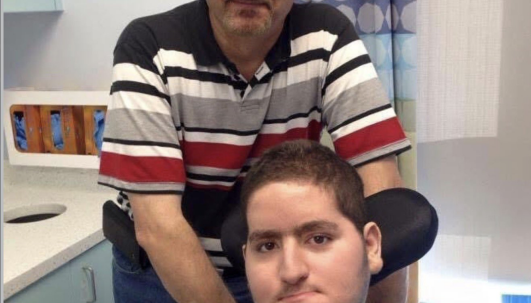 Wael Tarabishi, a disabled man using a wheelchair and breathing tube, sits in a hospital room with his father and full-time carer, Maher Tarabishi, standing behind him.