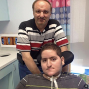 Wael Tarabishi, a disabled man using a wheelchair and breathing tube, sits in a hospital room with his father and full-time carer, Maher Tarabishi, standing behind him.