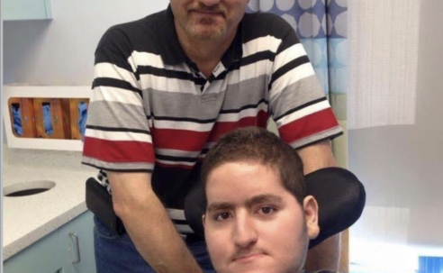 Wael Tarabishi, a disabled man using a wheelchair and breathing tube, sits in a hospital room with his father and full-time carer, Maher Tarabishi, standing behind him.