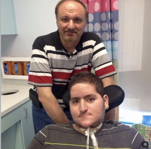 Wael Tarabishi, a disabled man using a wheelchair and breathing tube, sits in a hospital room with his father and full-time carer, Maher Tarabishi, standing behind him.