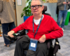 Colin Hughes wearing Ray-Ban Meta smart glasses and a red jumper, seated in his powered wheelchair at Meta’s AI Glasses Community Summit in London.