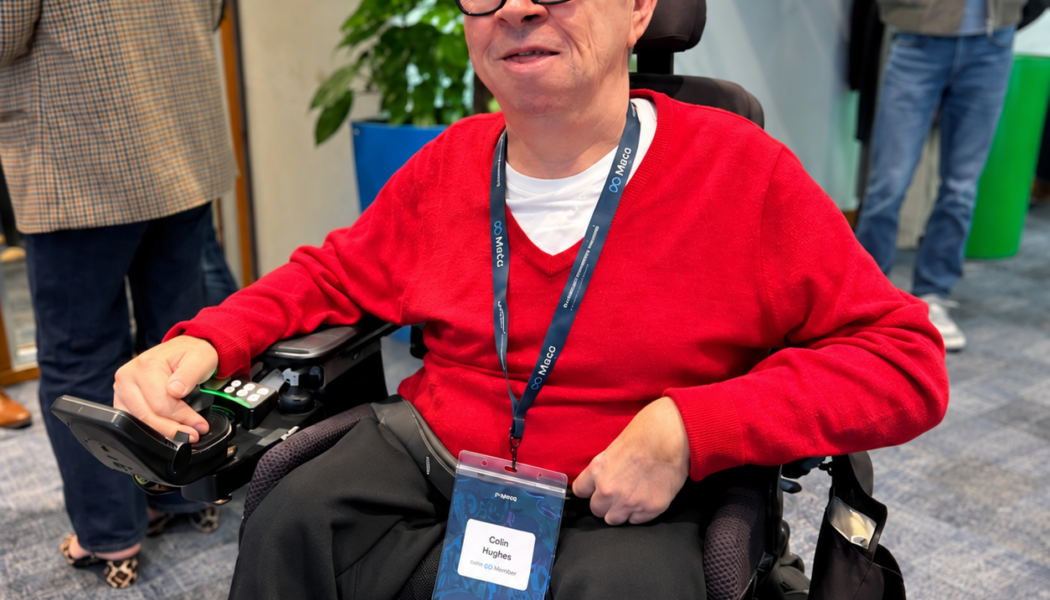 Colin Hughes wearing Ray-Ban Meta smart glasses and a red jumper, seated in his powered wheelchair at Meta’s AI Glasses Community Summit in London.