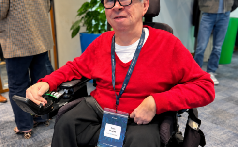 Colin Hughes wearing Ray-Ban Meta smart glasses and a red jumper, seated in his powered wheelchair at Meta’s AI Glasses Community Summit in London.