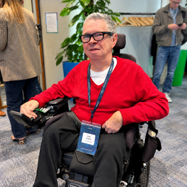 Colin Hughes wearing Ray-Ban Meta smart glasses and a red jumper, seated in his powered wheelchair at Meta’s AI Glasses Community Summit in London.