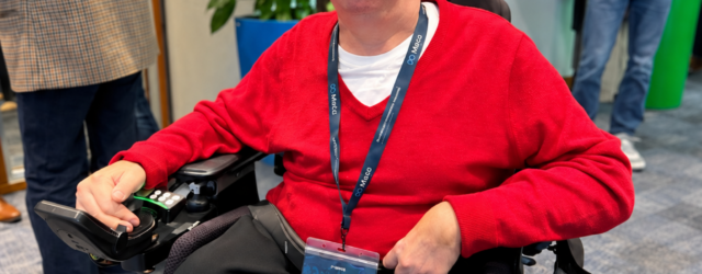 Colin Hughes wearing Ray-Ban Meta smart glasses and a red jumper, seated in his powered wheelchair at Meta’s AI Glasses Community Summit in London.