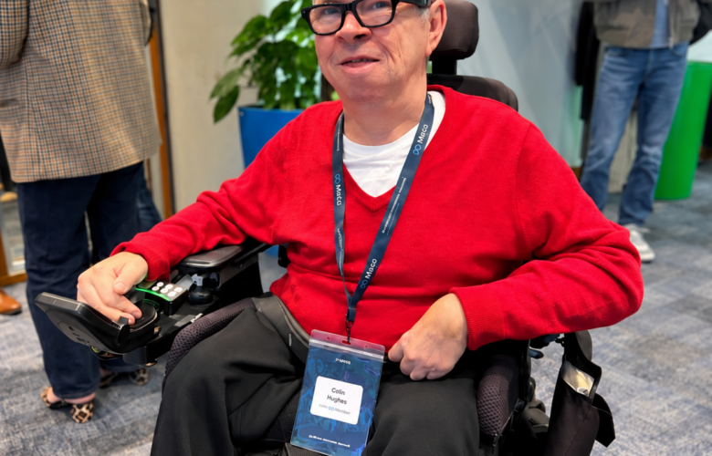 Colin Hughes wearing Ray-Ban Meta smart glasses and a red jumper, seated in his powered wheelchair at Meta’s AI Glasses Community Summit in London.
