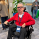 Colin Hughes wearing Ray-Ban Meta smart glasses and a red jumper, seated in his powered wheelchair at Meta’s AI Glasses Community Summit in London.